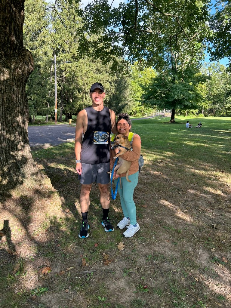 A man and woman, holding their dog, out in the park. Man has a race bib that says 148 on his shirt.