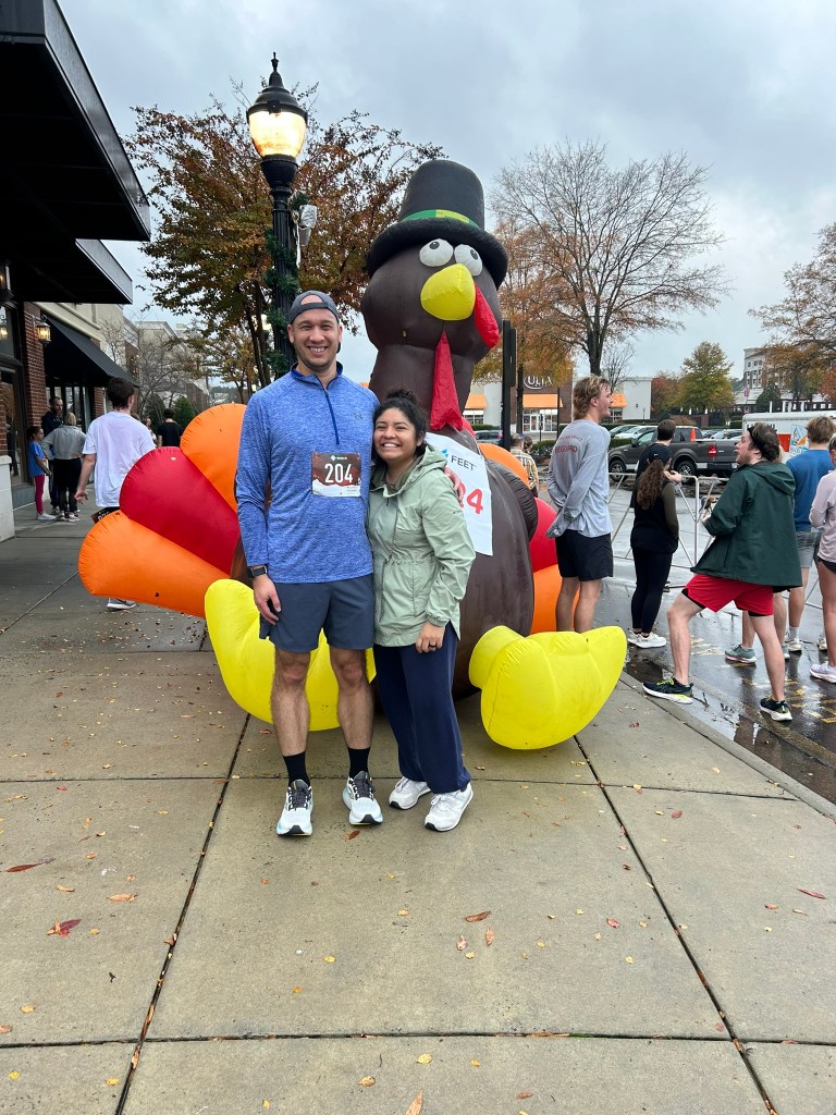 Man and woman, posing in front of an inflatable Thanksgiving turkey. People around as an event took place. Man has a race bib that says 204 on his shirt.