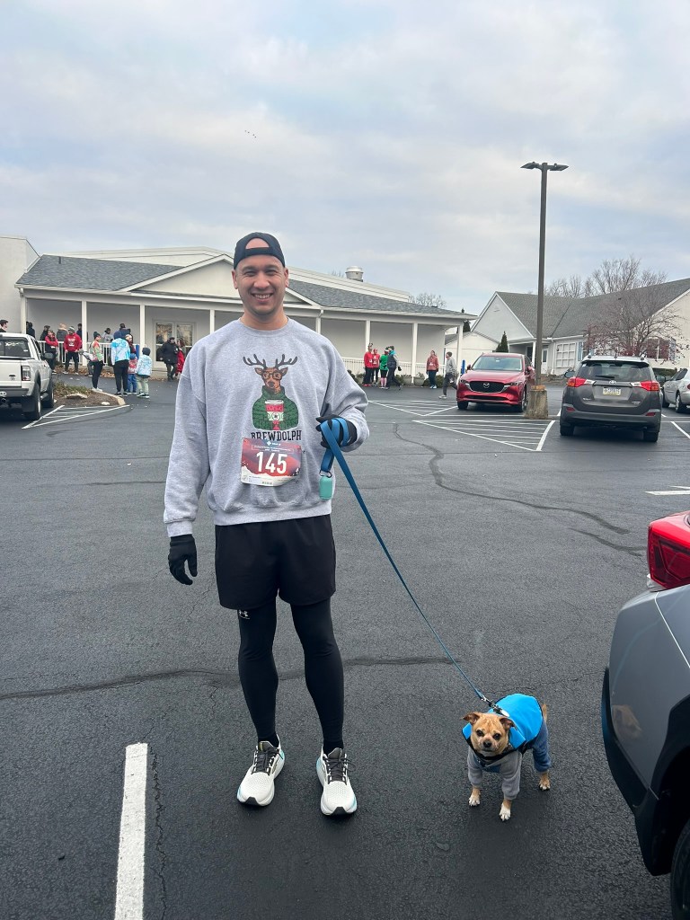 Man holding his dog on a leash in a parking lot, with a race bib that says 145 on his gray sweatshirt.
