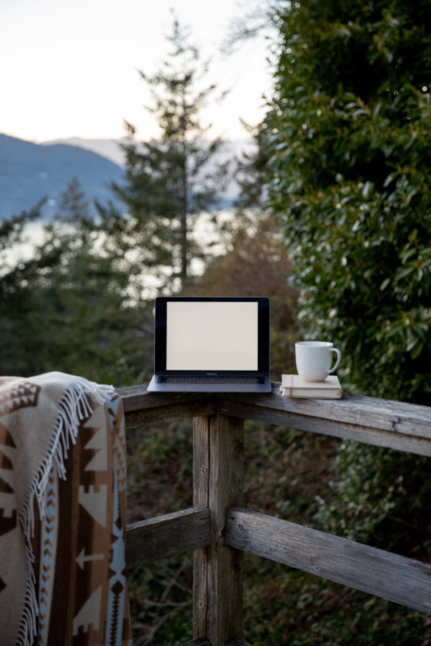 A laptop, sitting on the corner of an outdoor deck, with a mug on top of a journal. A blanket is draping over the railing of the deck on the left side of the laptop.