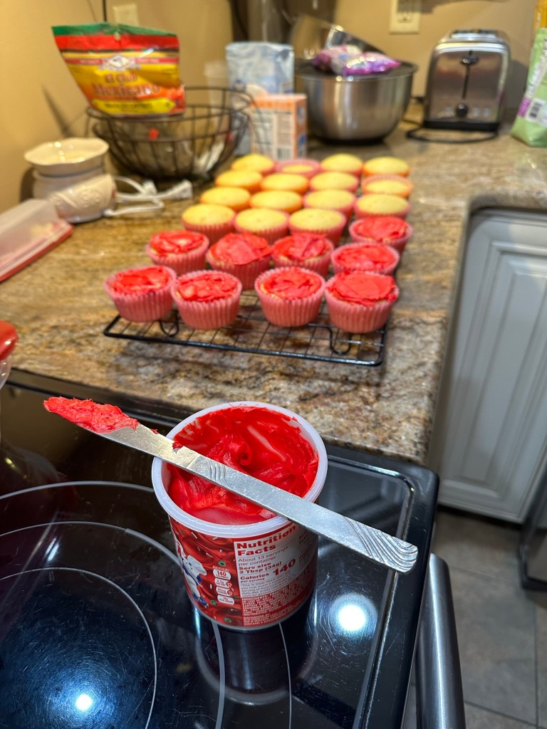 Vanilla cupcakes sitting on top of a drying rack. Half covered with red icing, while the rest wait for icing. The container of icing sits on top with a butter knife used to spread on top.
