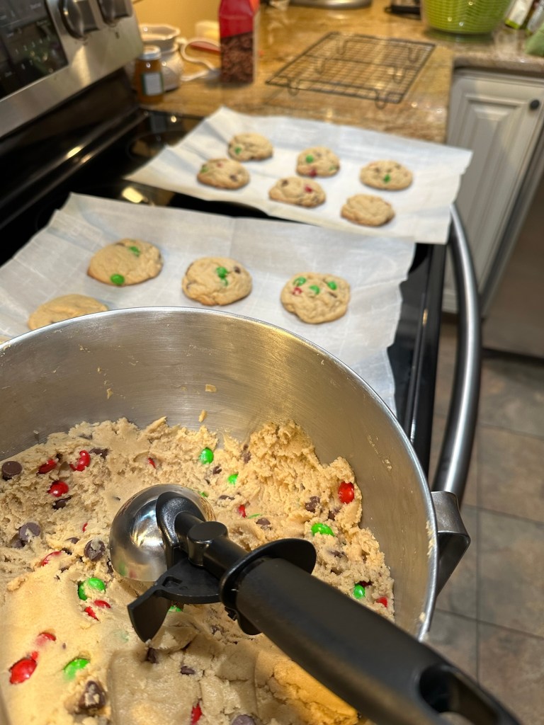 Mixing bowl of chocolate chip cookies dough, with a scooper inside. Baked cookies lie next to it on the cookie sheet after coming out of the oven.