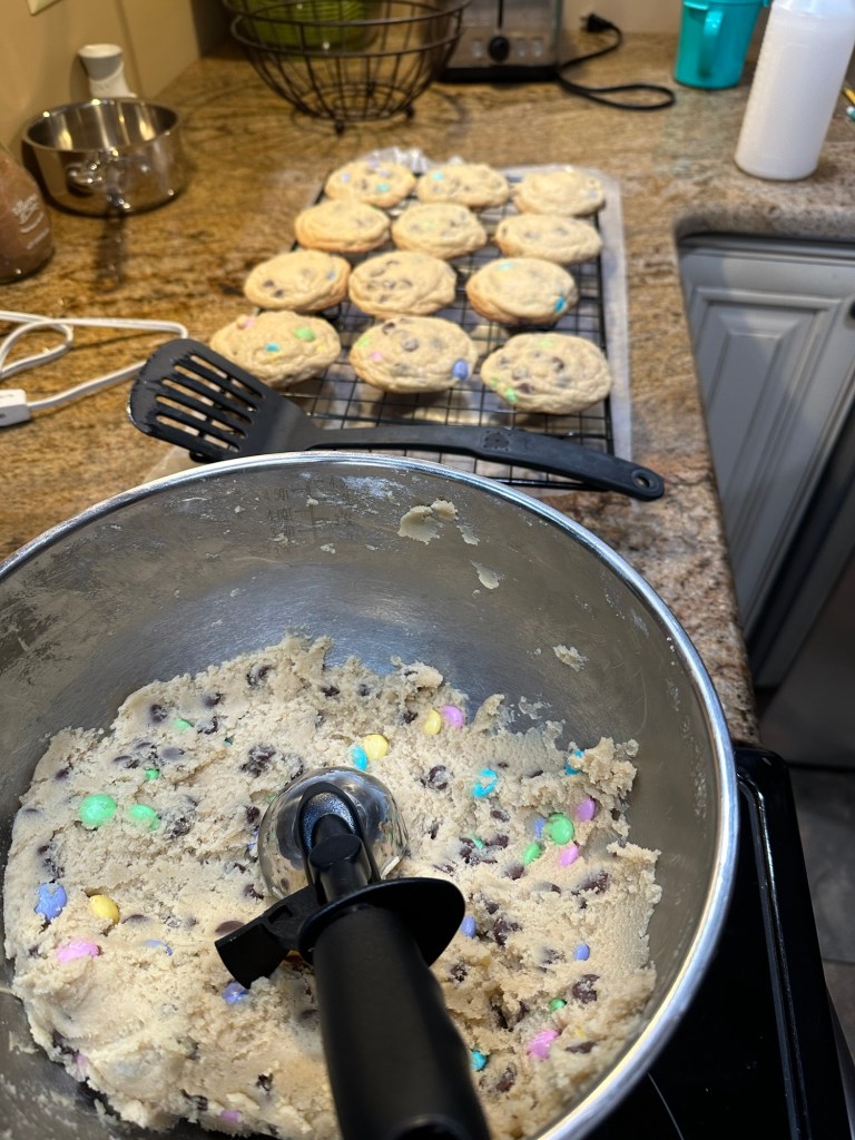 Mixing bowl full of chocolate chip cookies dough, with M&M’s added. Next is a flipped with already baked cookies, cooling on the drying rack.