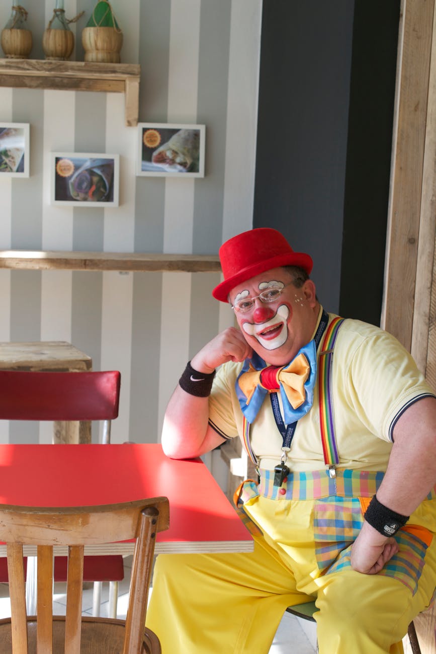 A clown, posing in front of the camera, with their arm resting their head on the table.